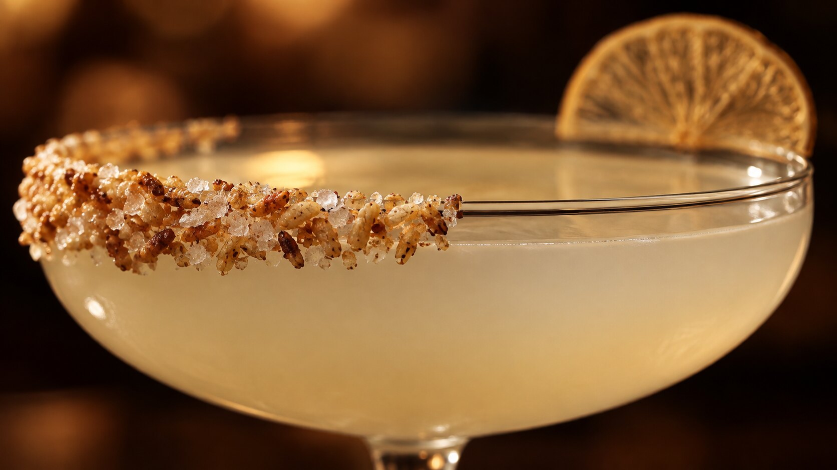 Primer plano extremo del borde de una copa coupe con una costra de arroz tostado y sal de grano de un solo lado, y una rodaja de c&iacute;trico deshidratado balanceada en el borde opuesto, la bebida p&aacute;lida brillando con luz de bar.
